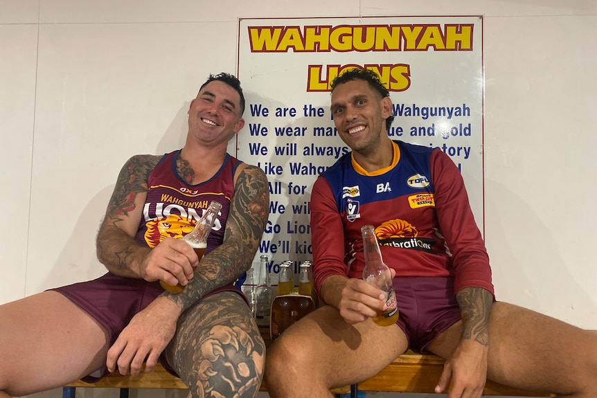 Two men wearing maroon football jumpers smile holding a beer each