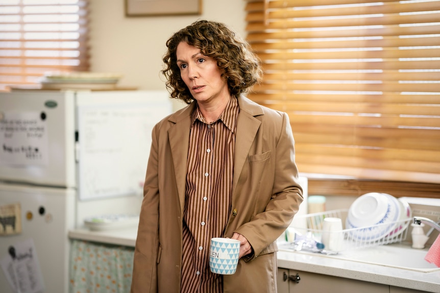Actor Kitty Flanagan as Helen-Tudor Fisk, wearing a brown suit and holding a blue and white mug.