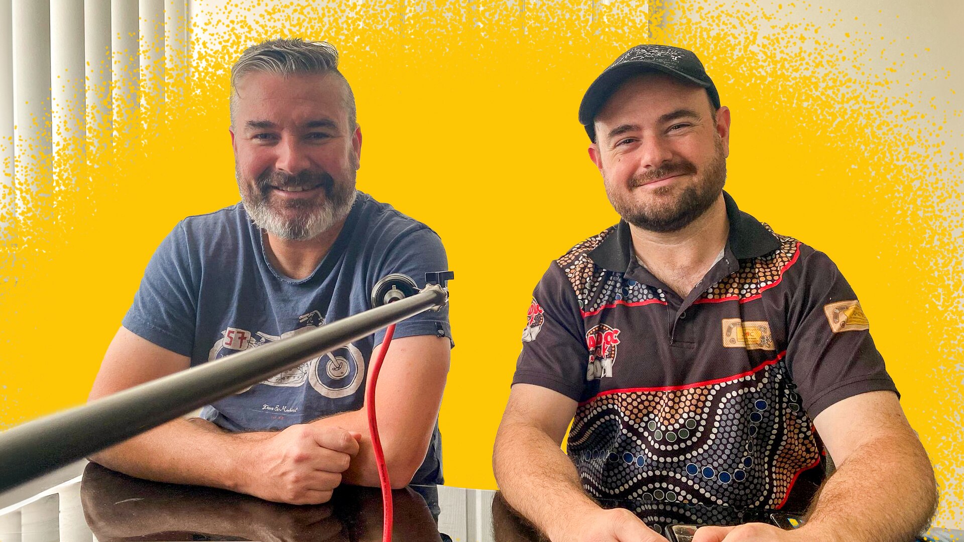 A man in his early 40s with short, salt and pepper hair and beard sits next to another man wearing a maroon short and cap. 