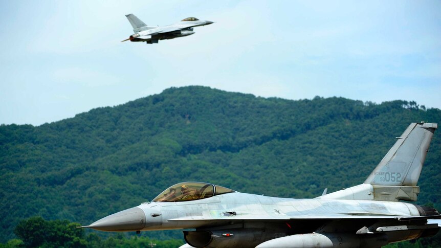 A Korean F-16 fighter jet is on a runway with green hills behind it while another jet is pictured in the distance in flight.