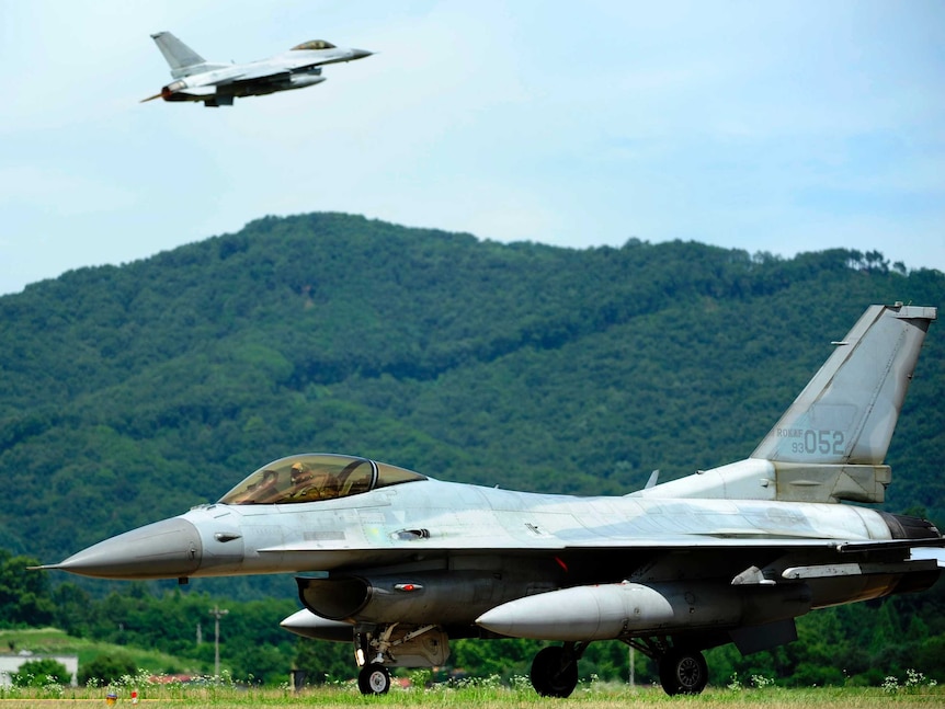 A Korean F-16 fighter jet is on a runway with green hills behind it while another jet is pictured in the distance in flight.