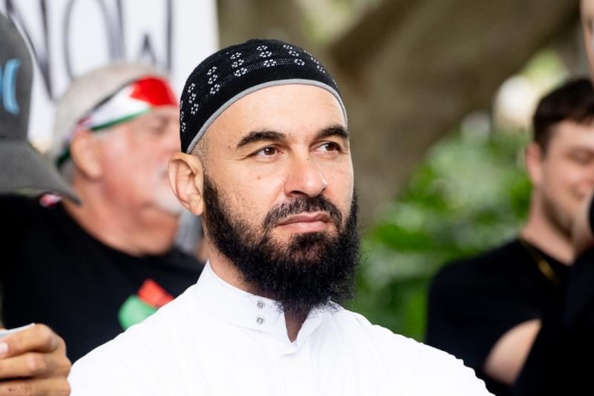 A muslim man in a white top looks at the camera, holding a jacket