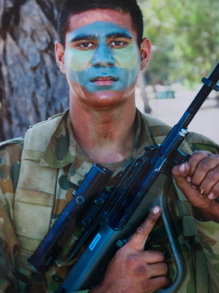Daniel Dawson with blue and green paint on his face. Wearing army uniform, holding a weapon.