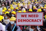 Protesters wearing yellow hard hats sit, one holds placard reading 'we need democracy'