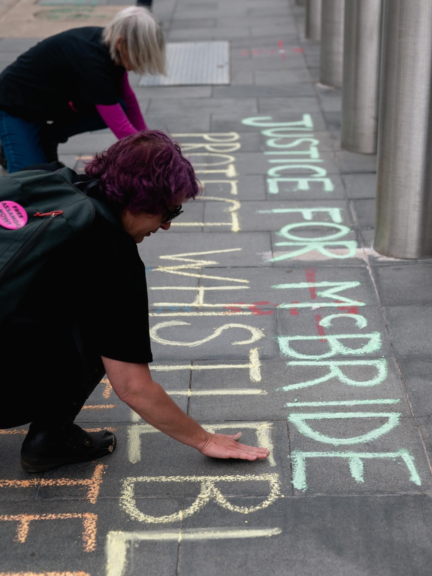 Two people courch to write 'Justice for McBride Protect Whistleblowers' in coloured chalk on tiled footpath.