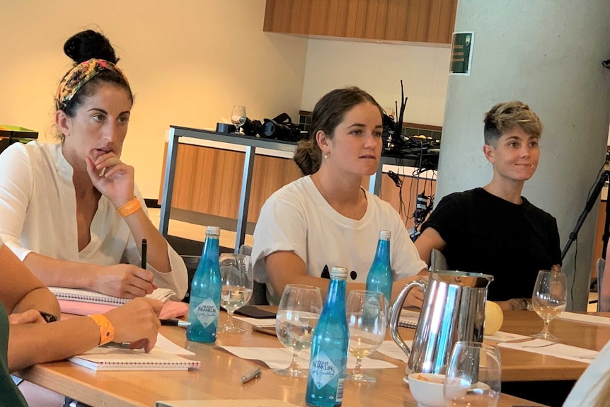 Four elite women athletes sit at a table and listen to their teacher.