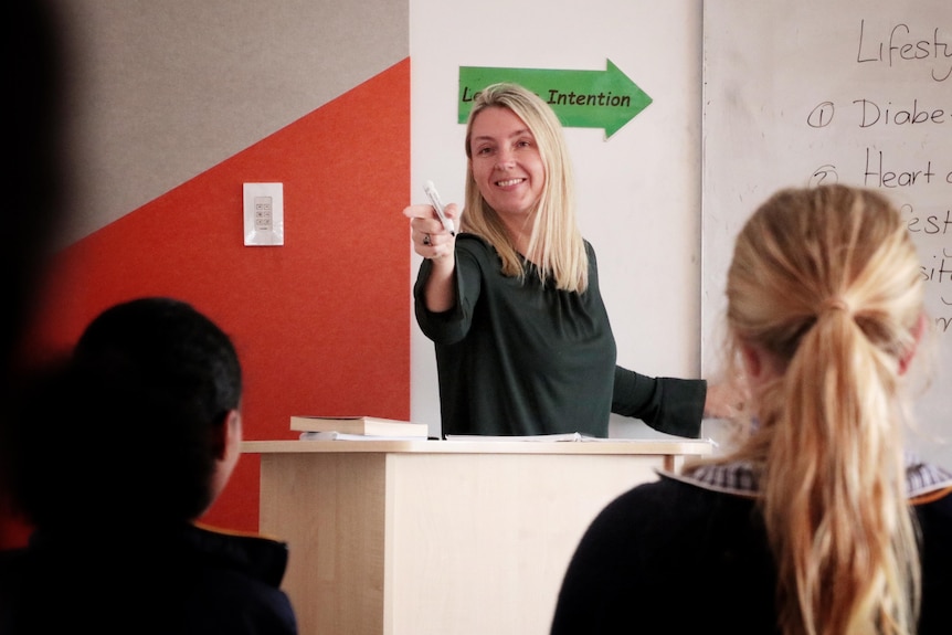 A teacher with blonde hair speaks to students next to a whiteboard.