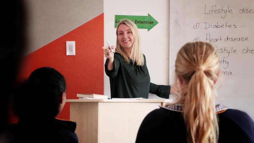 A teacher with blonde hair speaks to students next to a whiteboard.