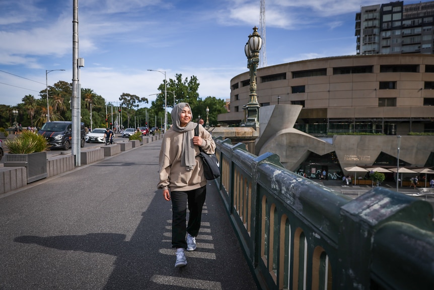 Adiba walks across a bridge across the Yarra River with the Arts Centre in the background