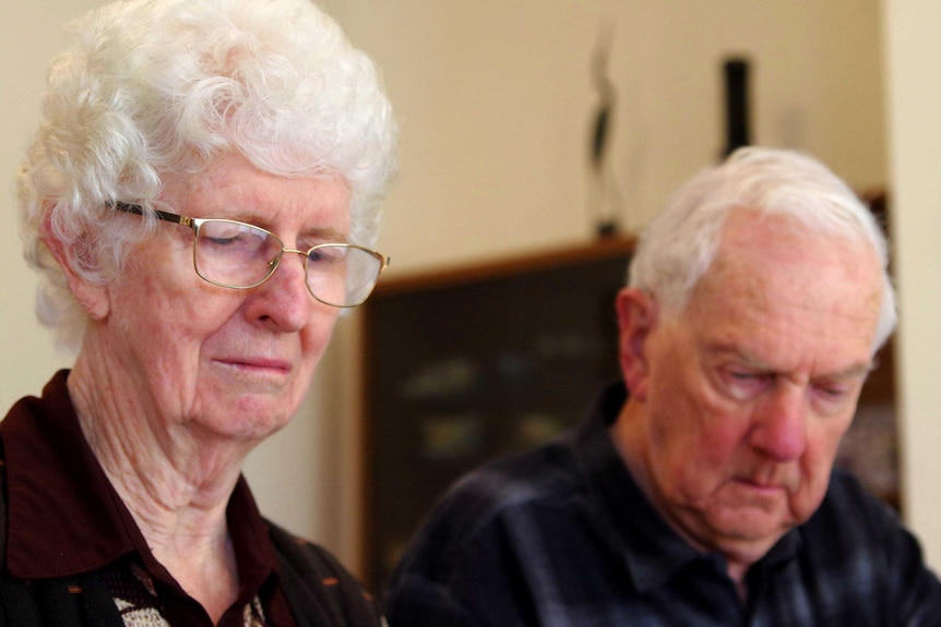 An elderly woman wearing glasses and her husband sit at a kitchen table.