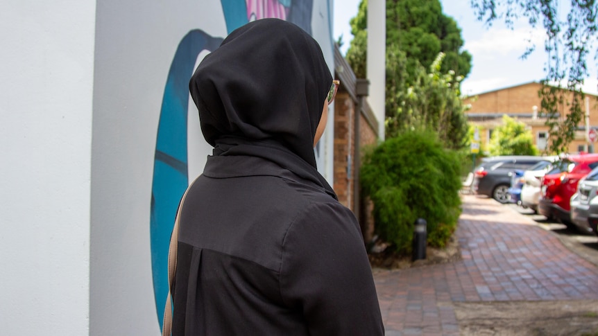 A young woman with a black hijab looks away from the camera, parked cars are in the background