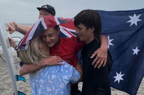 A teenage surfer with the Australian flag draped over him being carried by a woman and another teenager