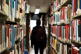 The silhouette of a young man wearing a backpack can be seen between the aisles of a library.