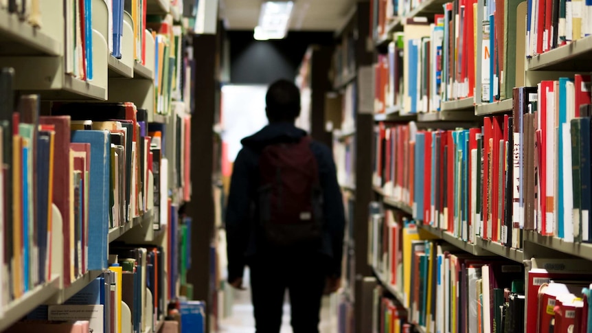 The silhouette of a young man wearing a backpack can be seen between the aisles of a library.