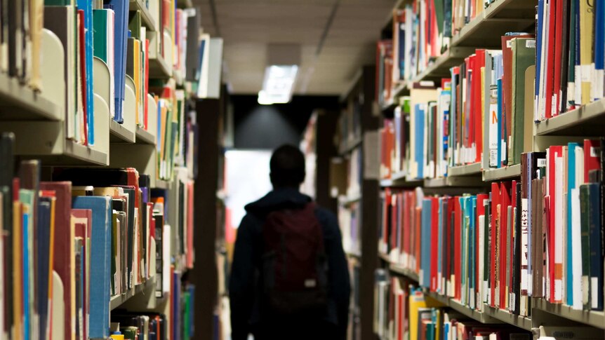 A child wearing a backpack walking between two book shelves at a school library.