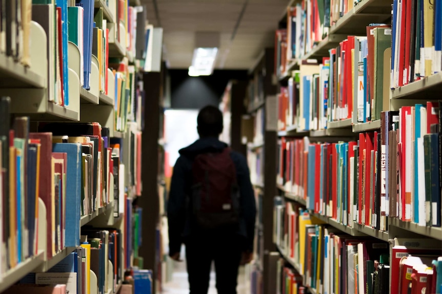 A child wearing a backpack walking between two book shelves at a school library.