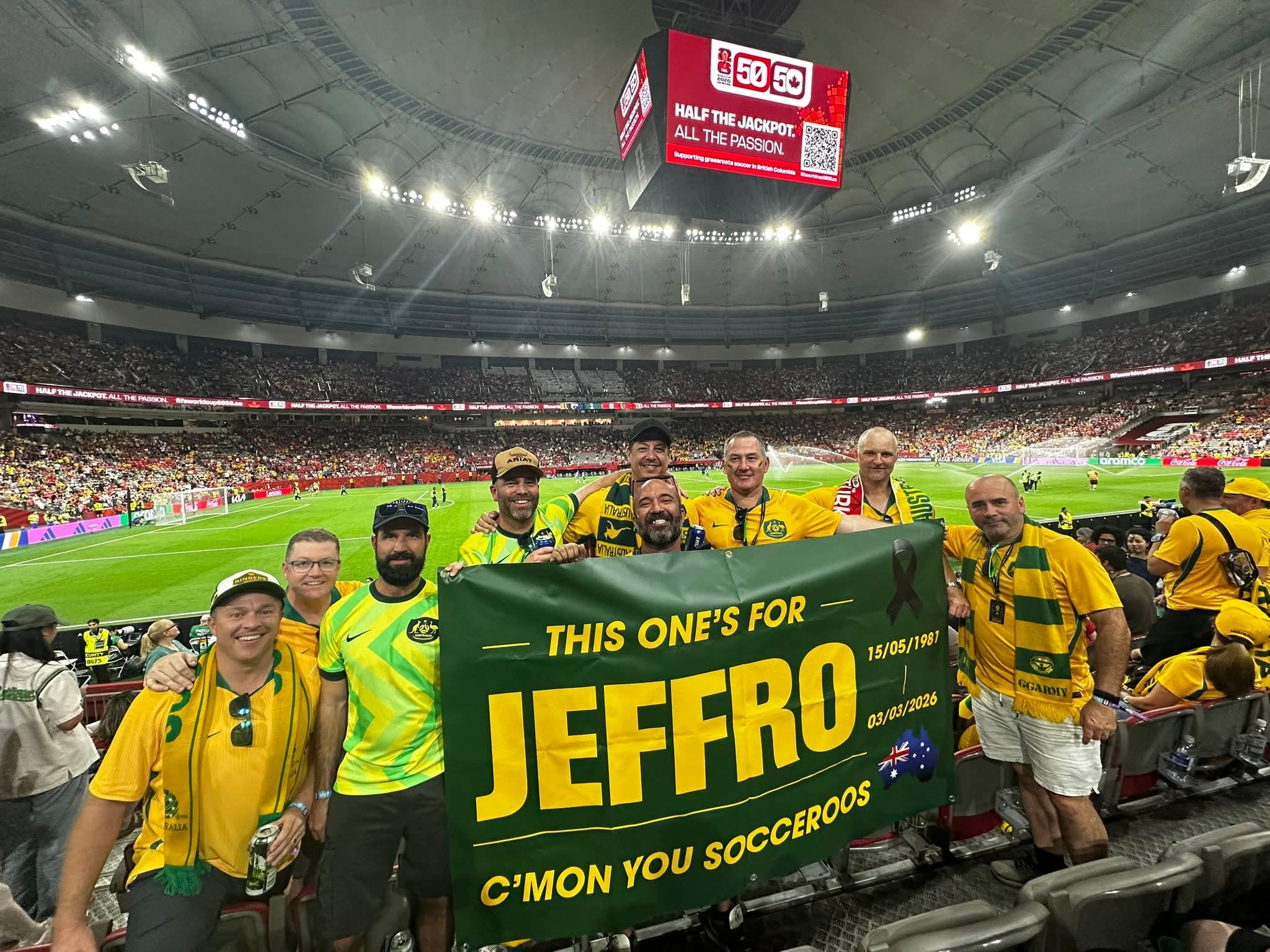 A group of men in jerseys in a sports stadium hold up a banner commemorating a late friend.