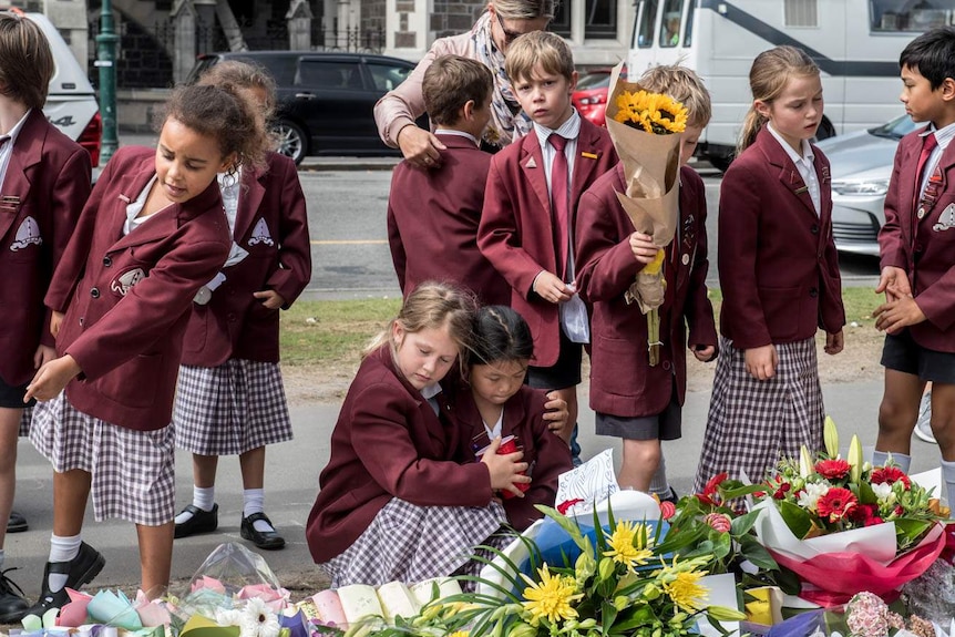 Two schoolgirls embrace, surrounded by their friends, while others lay flowers