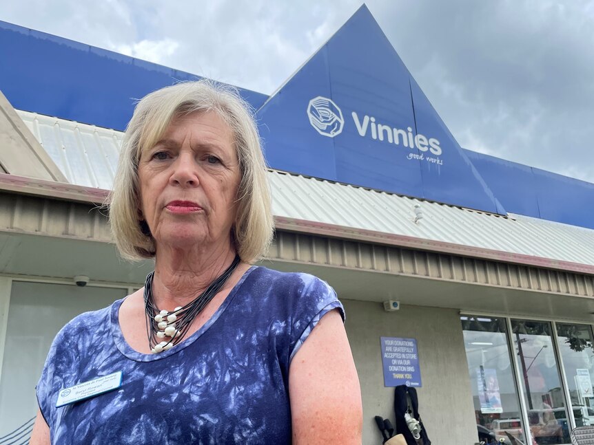 Una mujer con cabello rubio hasta los hombros, vestida con una blusa azul, parada afuera de una tienda Vinnies.