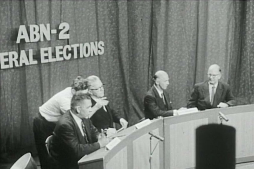 Black and white still of McMahon, Ward and two other guests sitting at panel with ABN 2 elections signage on curtain behind.