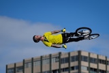 A rider on a BMX bike is high in the sky and on his side, with an apartment building in the background. 