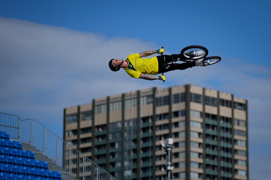 A rider on a BMX bike is high in the sky and on his side, with an apartment building in the background. 