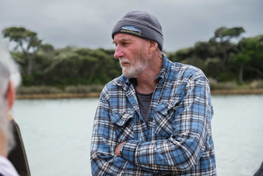 A 50-something man with a white beard, checked shirt and beanie folds his arms and listens with a river behind him