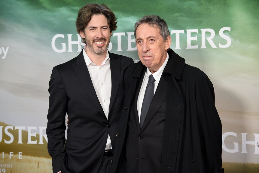 A young man stands next to an older man in front of a media wall with the word Ghostbusters.