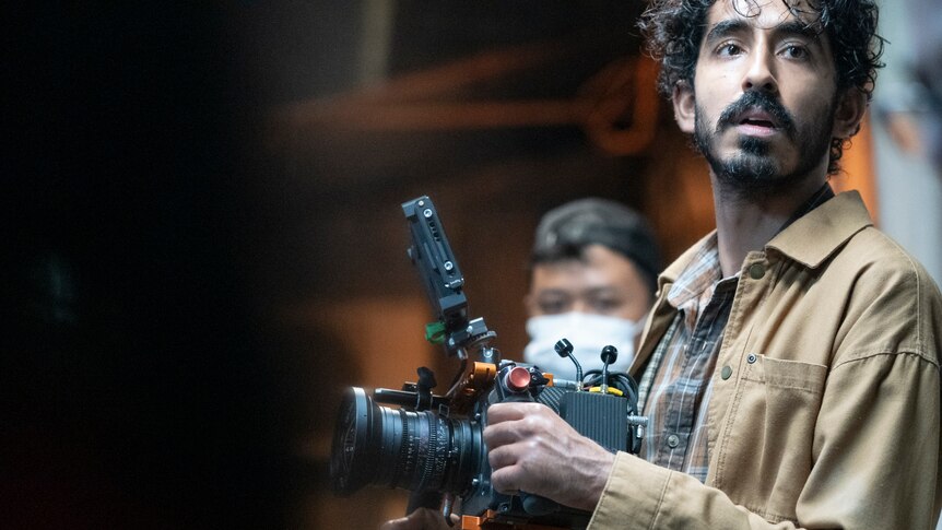 A man of South Asian descent with black hair and a moustache with a large camera on a film set