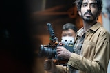 A man of South Asian descent with black hair and a moustache with a large camera on a film set