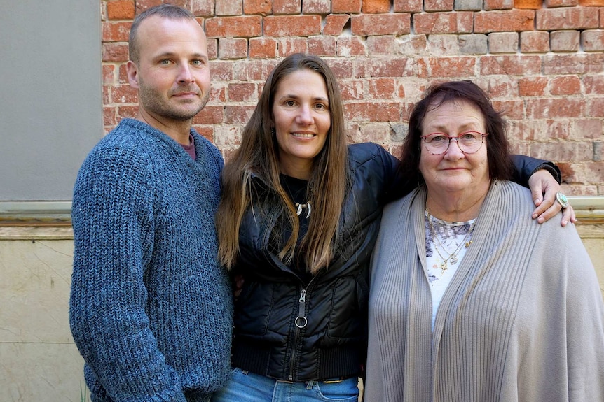 A man, and two women embrace in front of a brick wall.