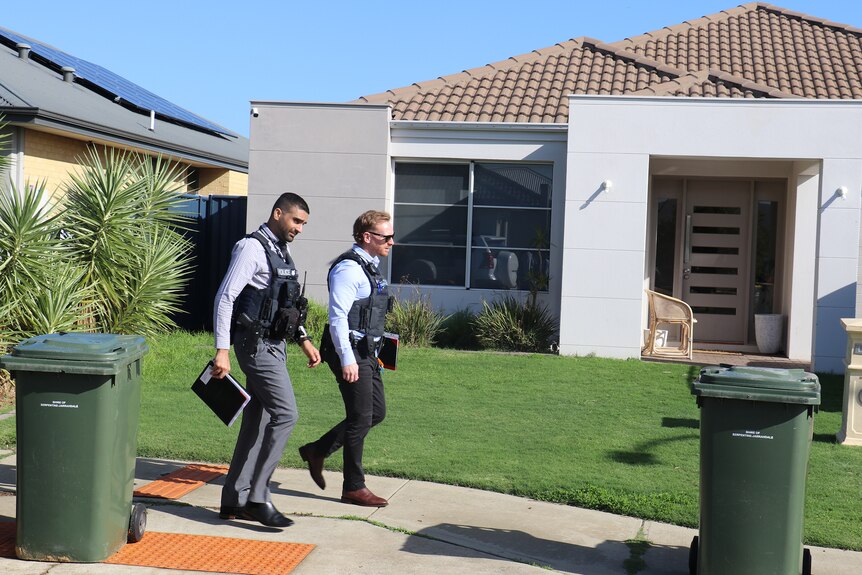 Two plain-clothes male police officers wearing bullet-proof vests walk along a footpath in front of a house.