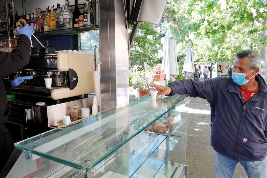 A man wearing a mask reaches out to take a small takeaway coffee from a bench at a coffee stand in a park
