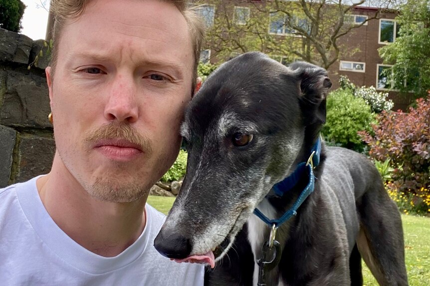 Patrick Lenton is wearing a white-t-shirt and posing for a selfie with his dog Basil, a black greyhound