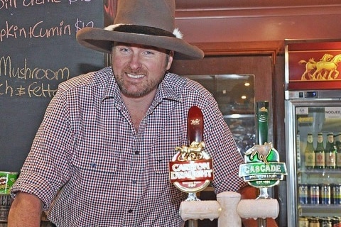 Simon stands behind the bar wearing a large hat and checked shirt 