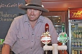 Simon stands behind the bar wearing a large hat and checked shirt 