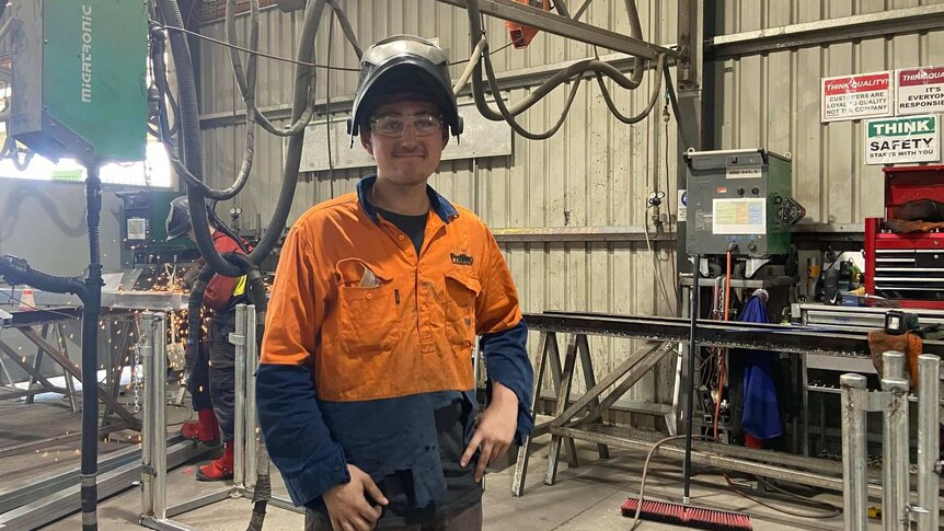 A young man in a high-vis shirt stands in front of welding equipment.