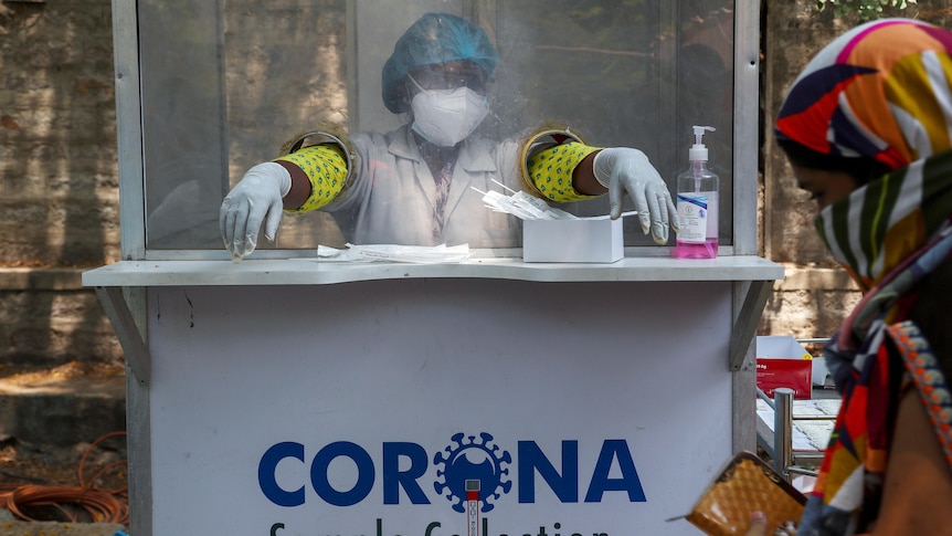 A masked health worker sits in a shielded booth with gloved hands sticking through a perspex protective screen.