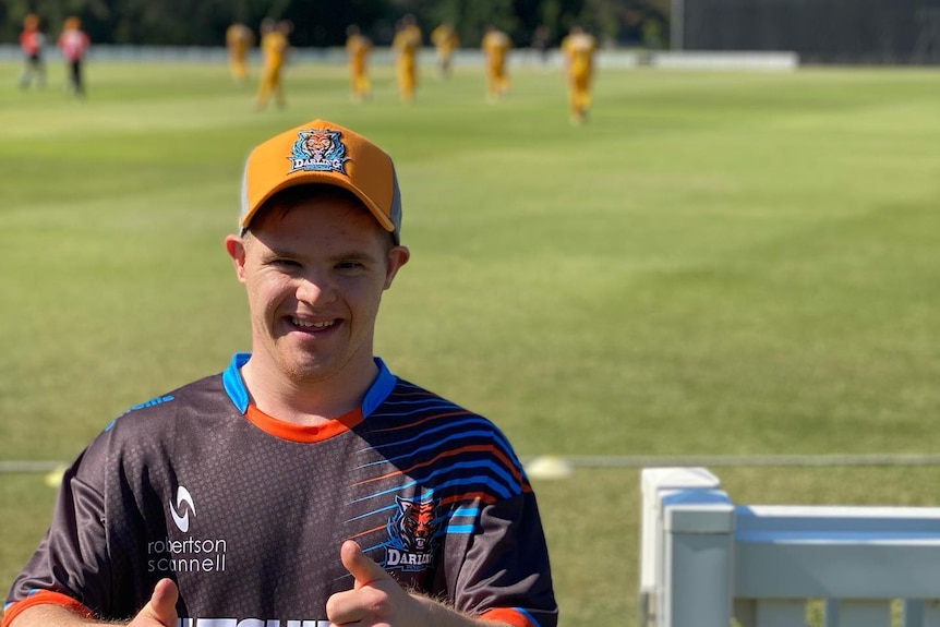 A man with a cap on smiling at the camera and cricketers in the background. 