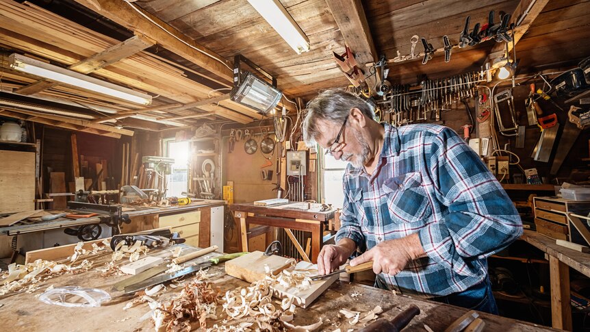 An middle-aged man in a flannel shirt chisels a piece of wood in his shed.
