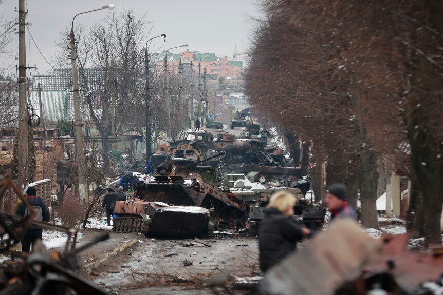 People mill around a long column of detroyed vehicles on a road in Ukraine