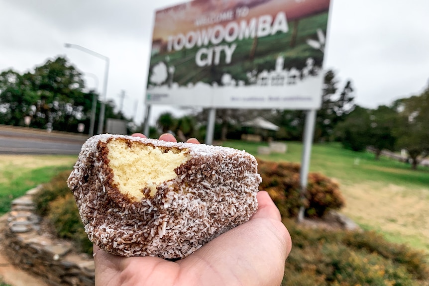 A lamington being held up in front of a 'Welcome to Toowoomba' sign