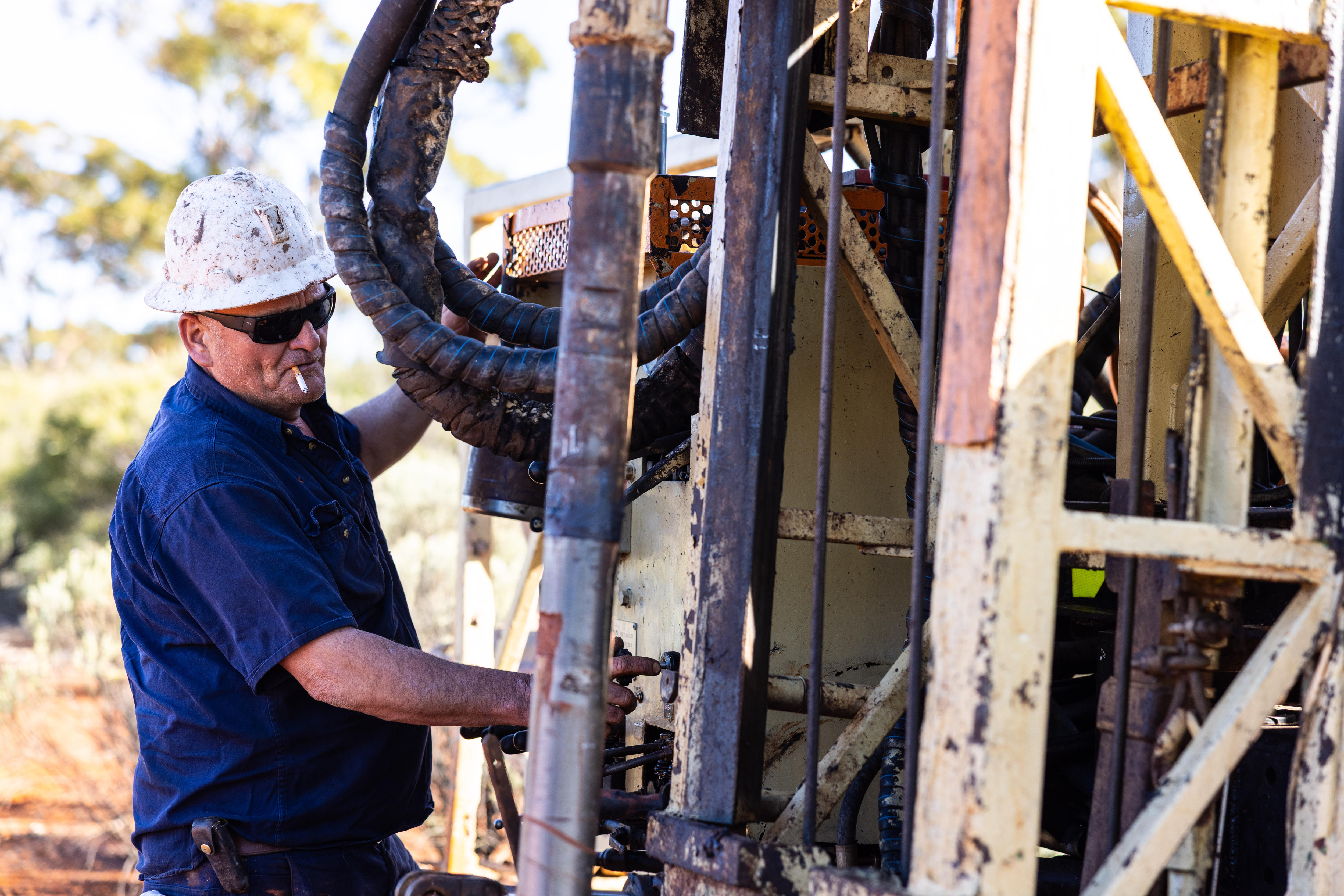 A man working controls of a drill rig in outback while wearing hard hat and smoking cigarette.  