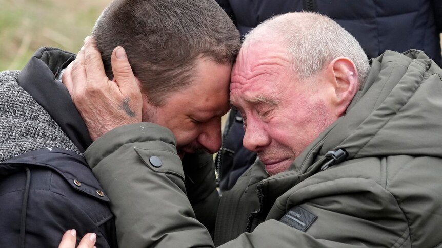 Two middle-aged men rest their foreheads against each other as they cry. 