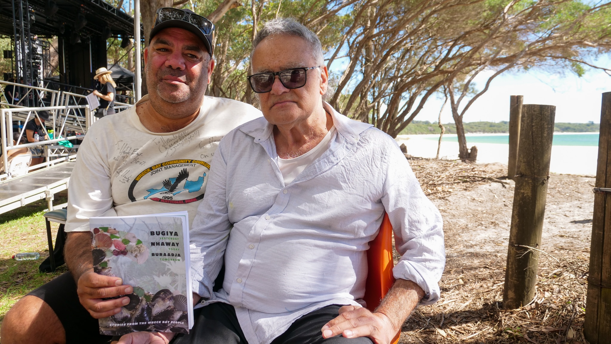 Двое мужчин сидят и держат книгу в мягкой обложке.