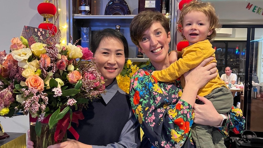 Two woman smiling and looking at the camera, one holding a bunch of flowers, the other a toddler.