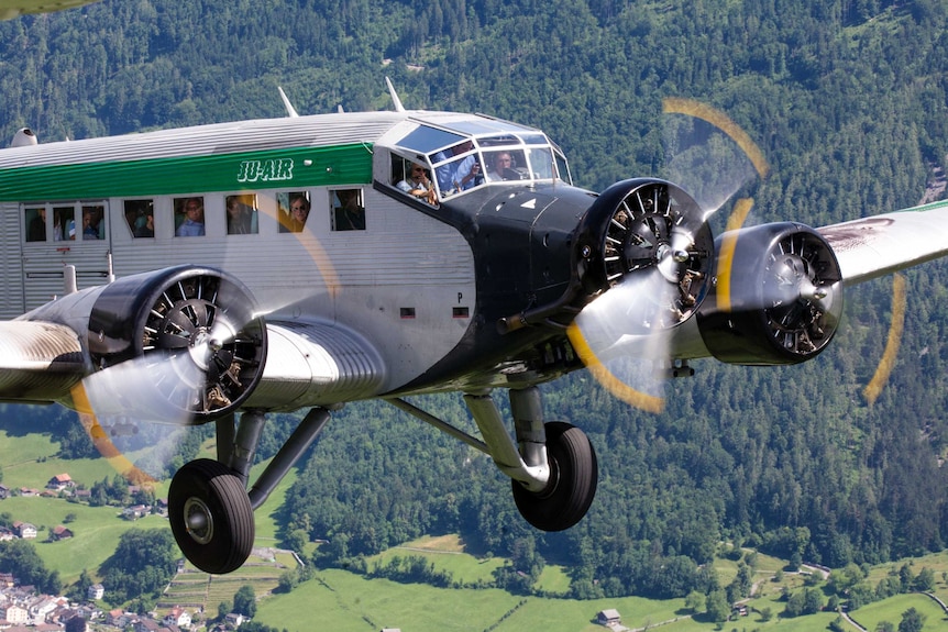 Passengers looking out of the JU-52 aircraft as it flies in the sky.