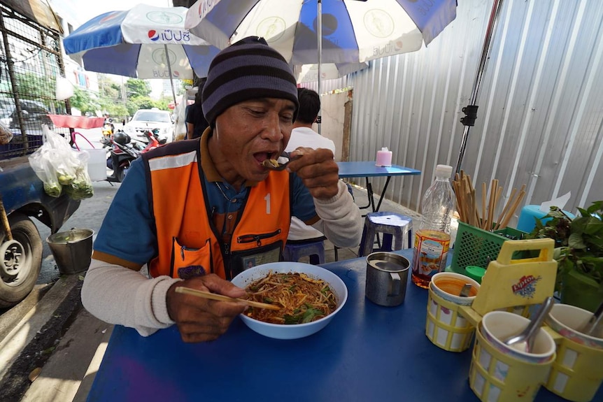 Motorcycle taxi driver eats noodles at a sidewalk eatery