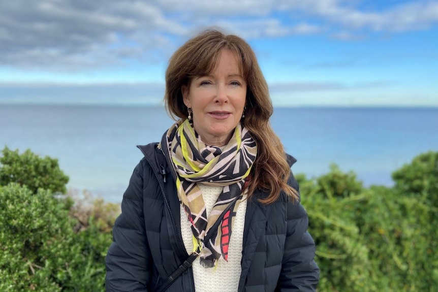 A woman with long brown hair wears a puffer jacket and a colourful scarf and stands in a park beside the ocean.