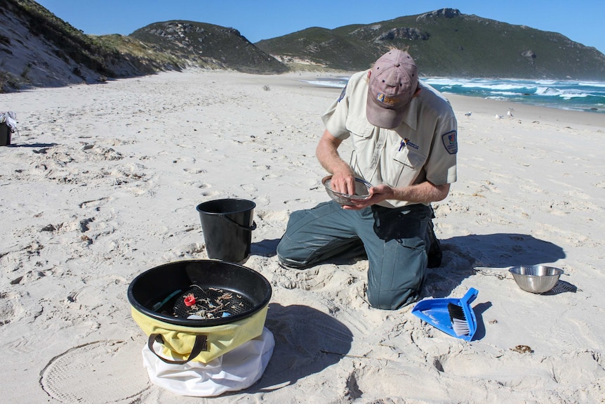 A man sits on a beach put sand through a spaghetti colander.